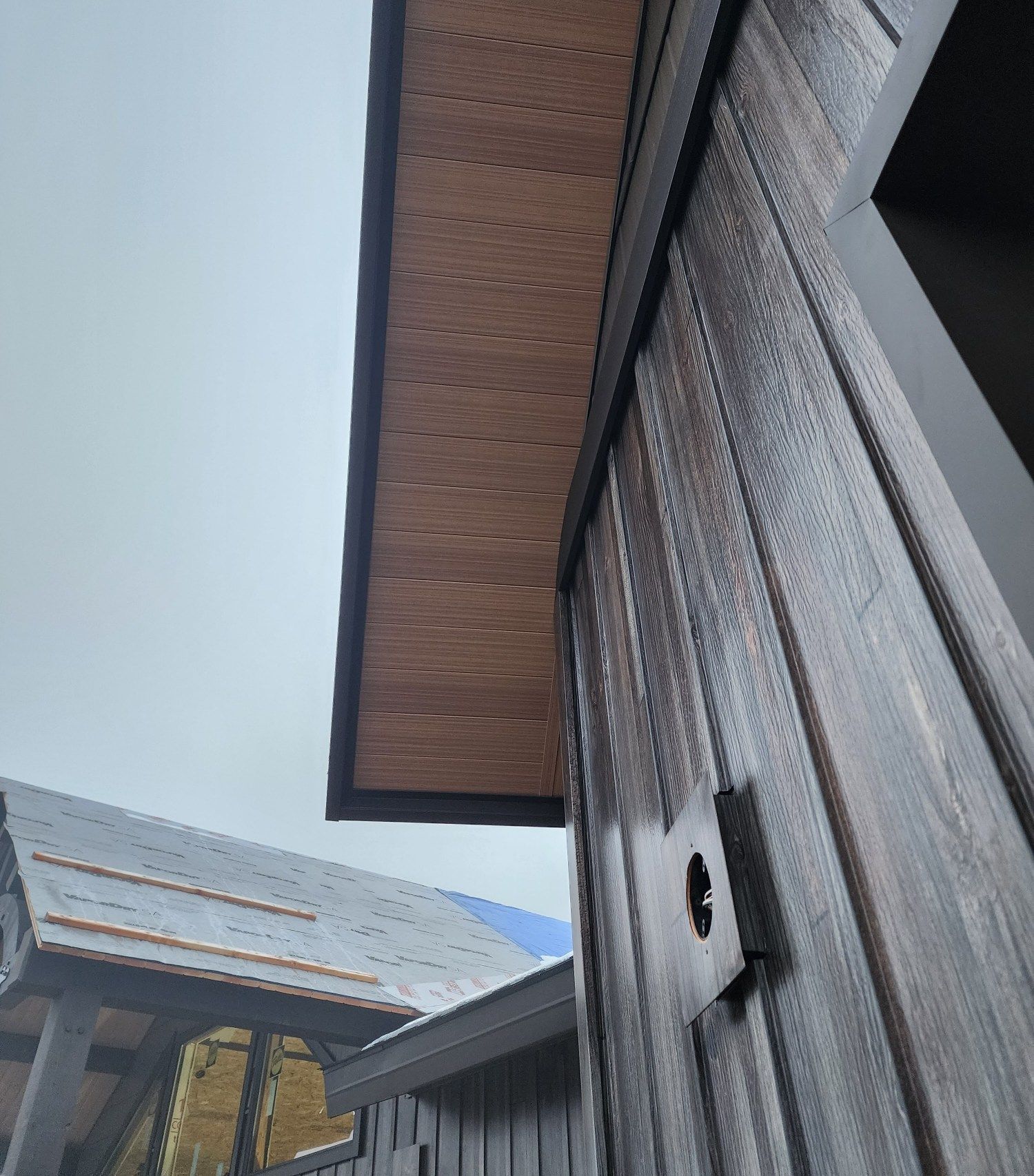 Exterior building detail with dark wood siding and a wooden overhang. Cloudy sky.