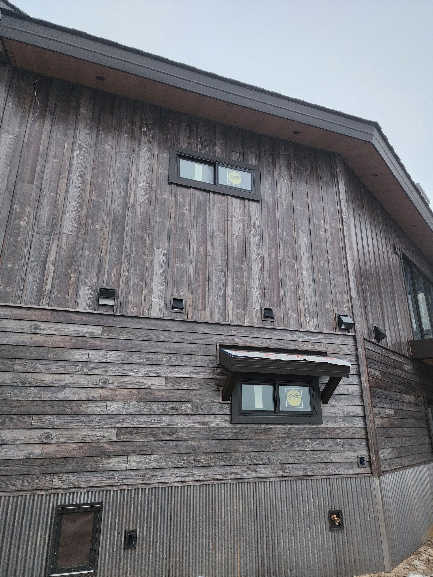 Exterior of a house with weathered wood siding and corrugated metal base. Windows have black frames.