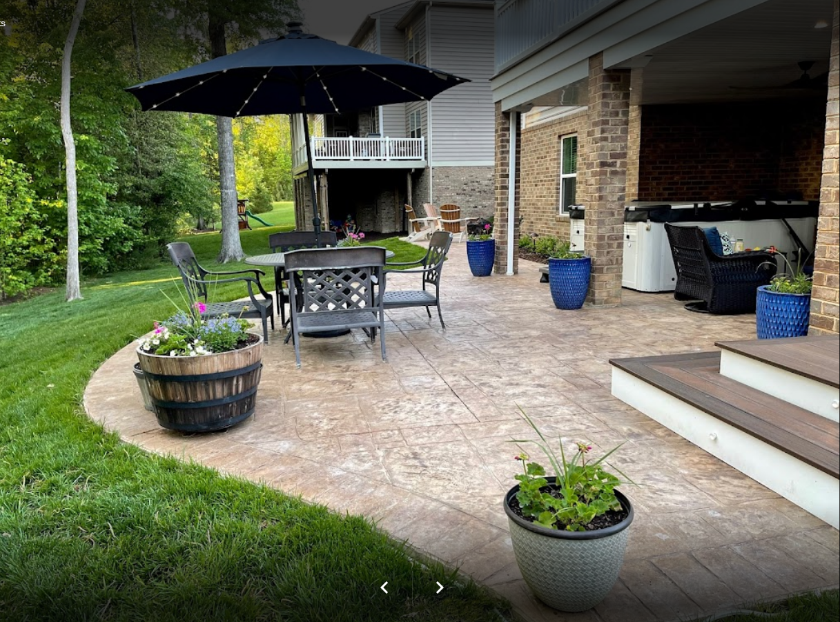 Patio with a black umbrella, table and chairs, and potted plants on a concrete surface.
