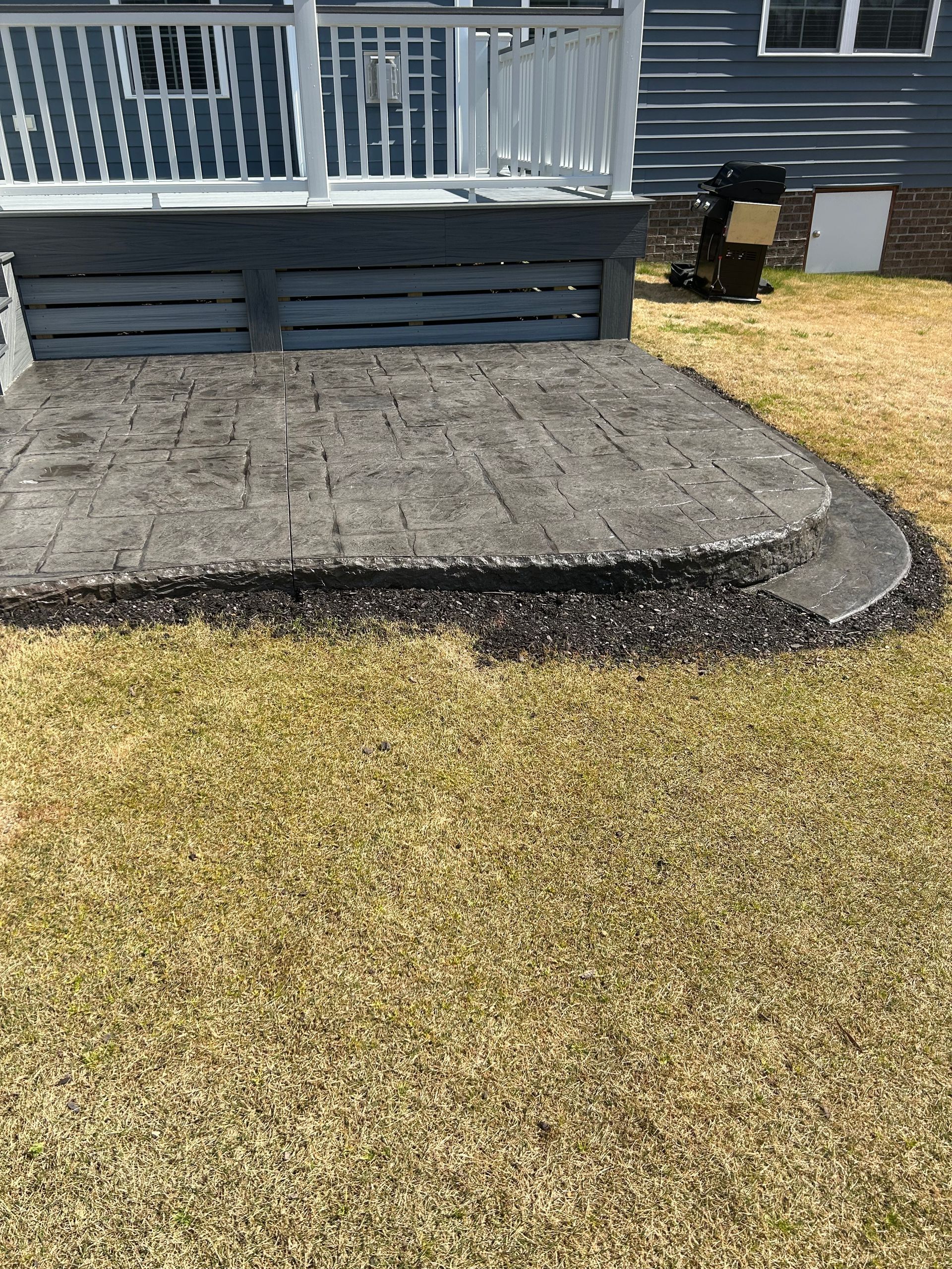 Stone patio with steps leading up to a deck, bordered by dark mulch and surrounded by brown grass.