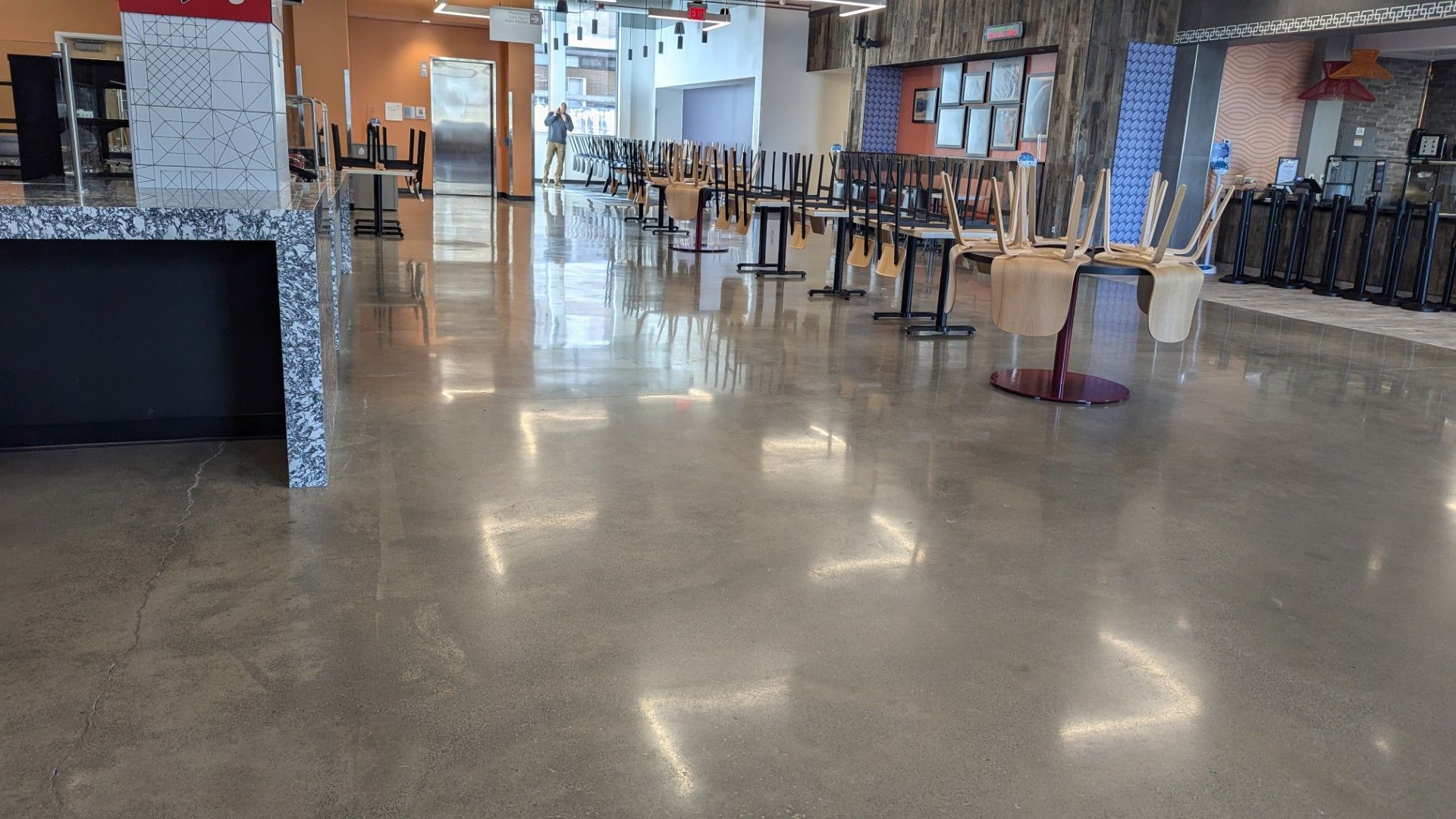 Polished concrete floor in an empty cafeteria with tables and chairs stacked. Bright lights reflect on the floor.