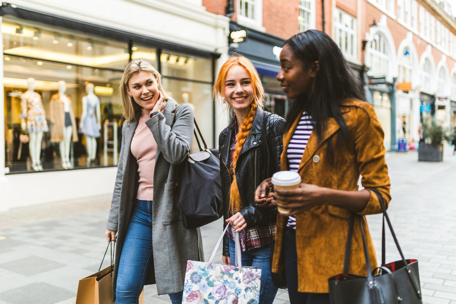 Three shoppers chatting on a city street, carrying bags and coffee near storefronts.