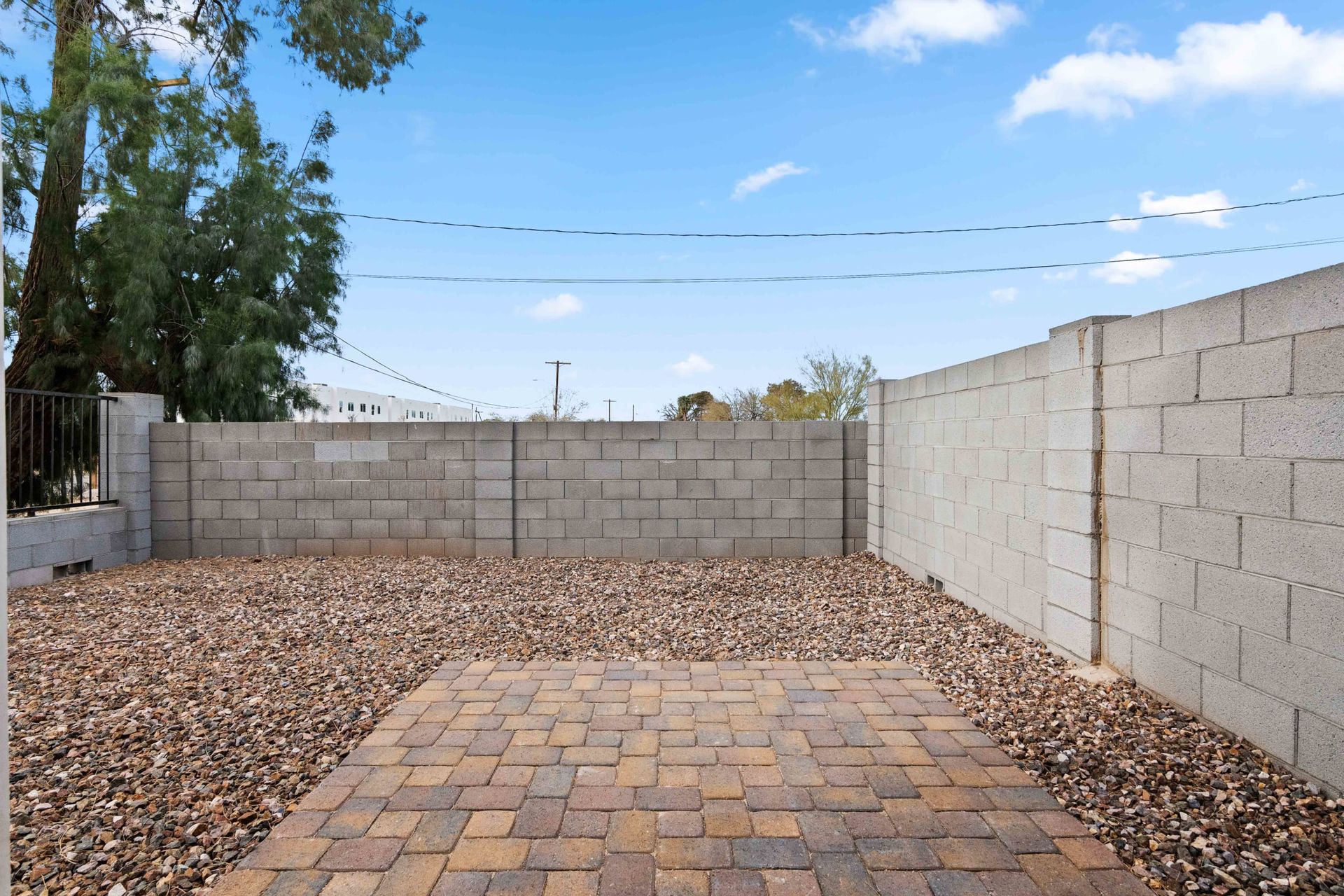 Small backyard with a brick patio, gravel ground, block walls, and a blue sky.