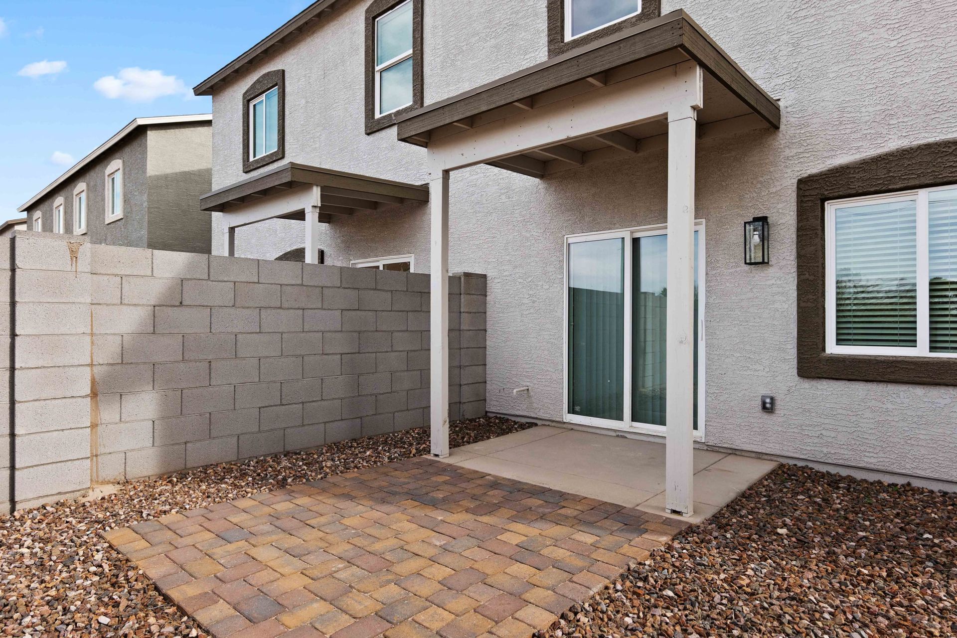 Covered backyard patio with brick pavers, stucco walls, sliding glass door, and white support posts