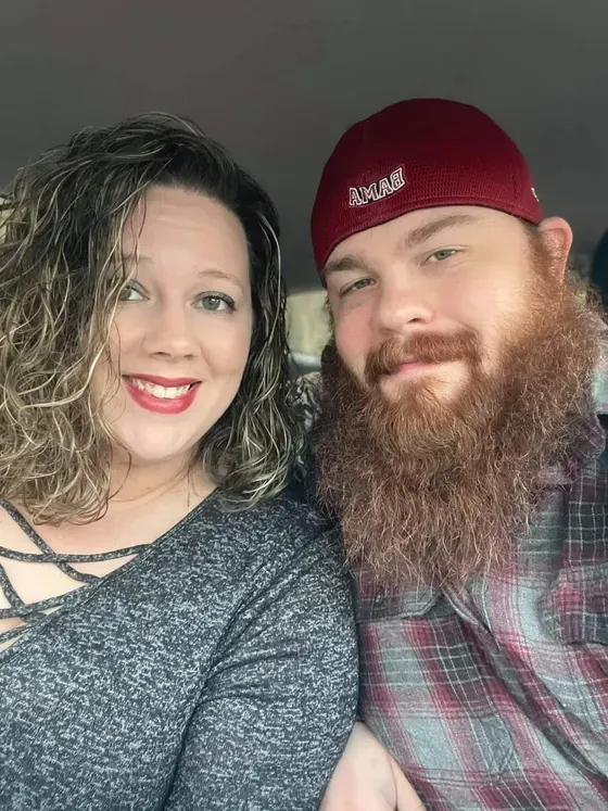 Woman with curly hair and red lipstick smiles next to a man with a large beard and a maroon cap.