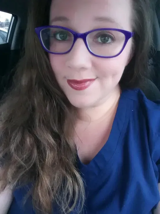 Woman with long brown hair wearing purple glasses and scrubs, smiling in a car.