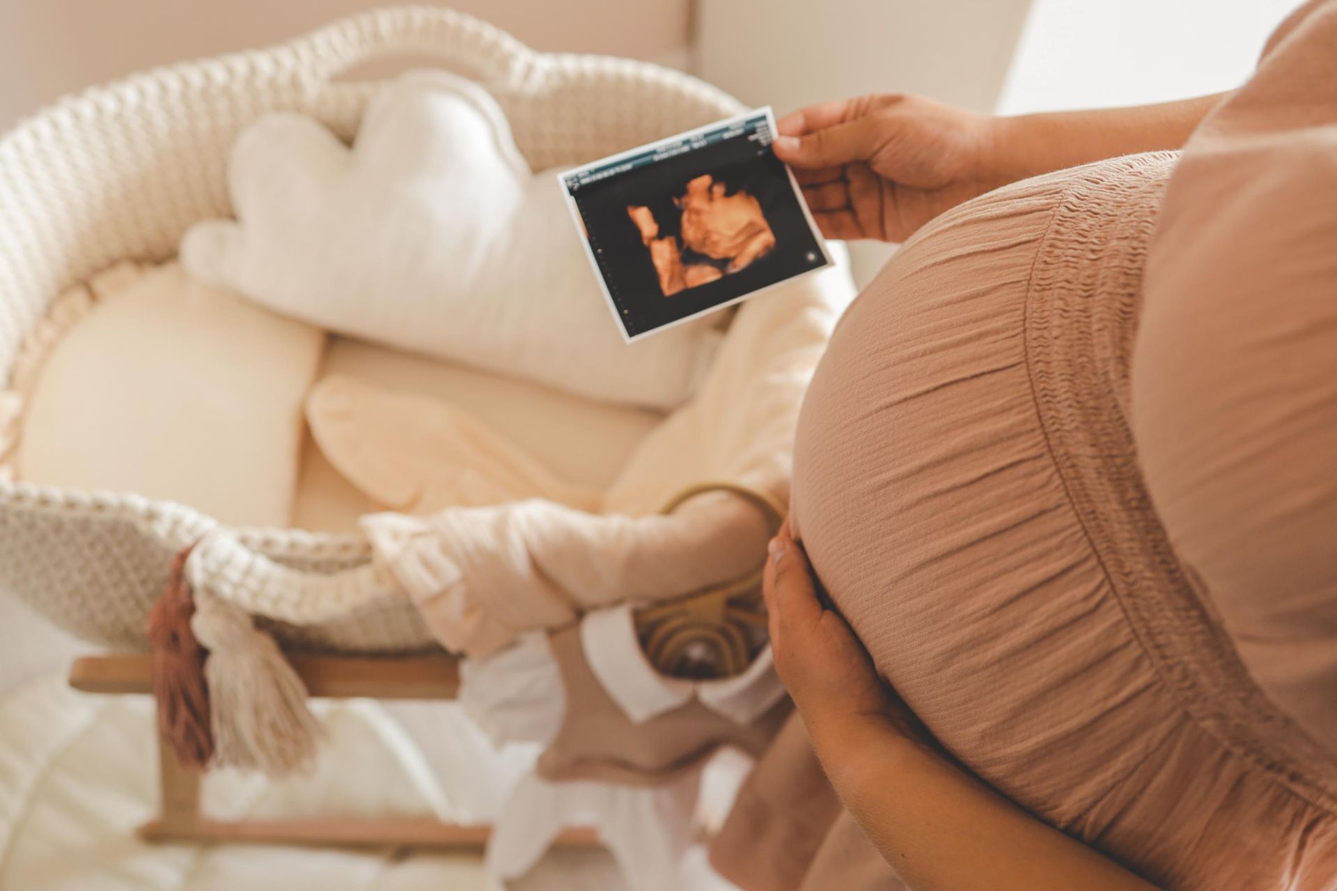 Pregnant woman holding ultrasound picture near a baby basket with soft toys and clothing.