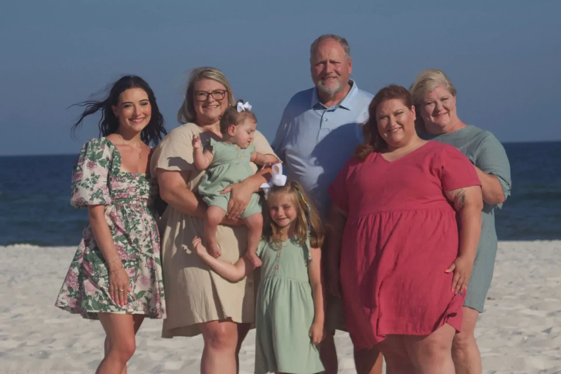 Family posing on a beach; seven people, adults and children; smiles; blue sky, white sand, ocean.