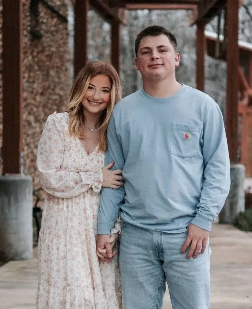 Couple smiling, holding hands, standing outdoors. Woman wears floral dress; man wears blue long sleeve shirt and jeans.