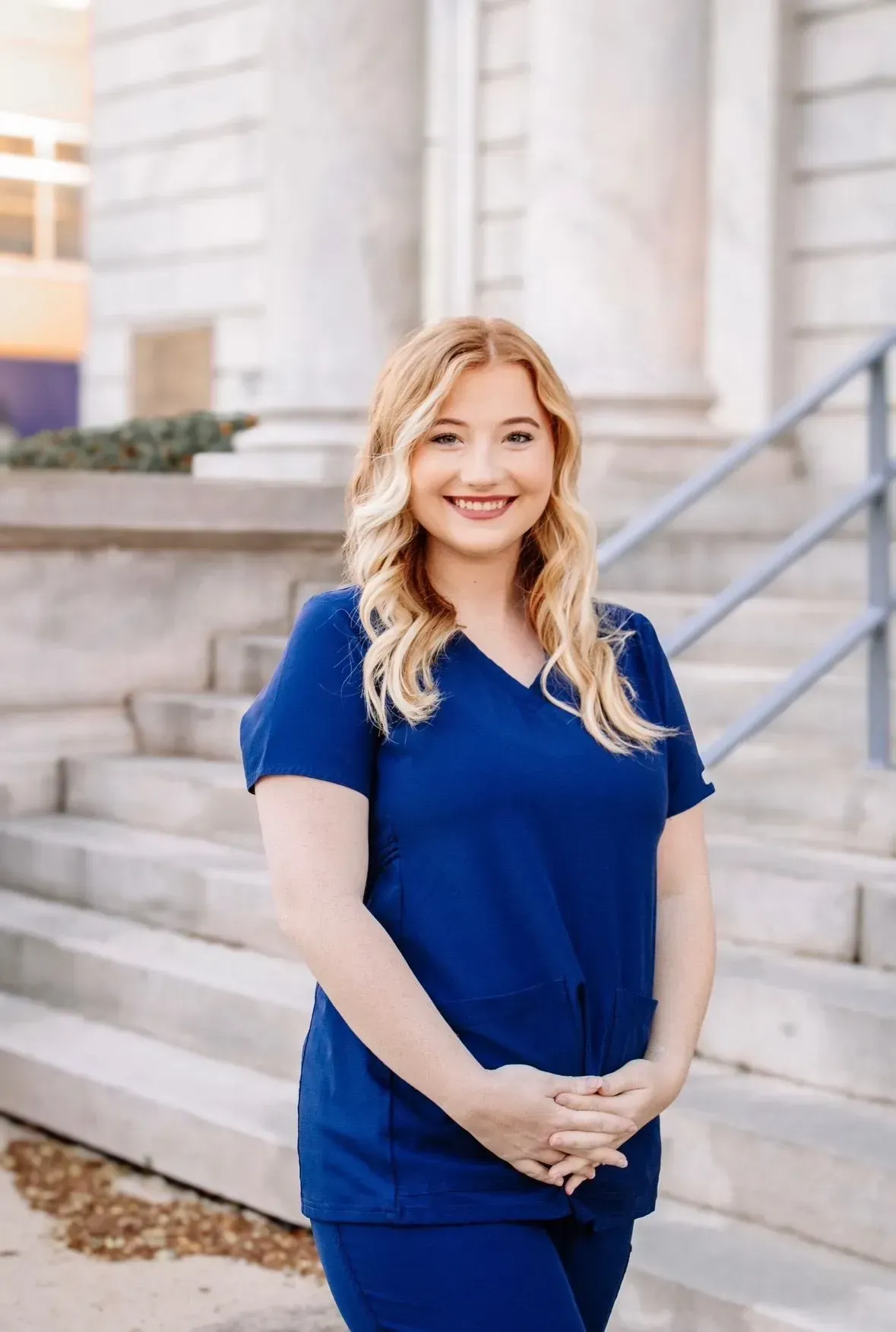 Woman with red hair in blue scrubs smiles on steps of a building.