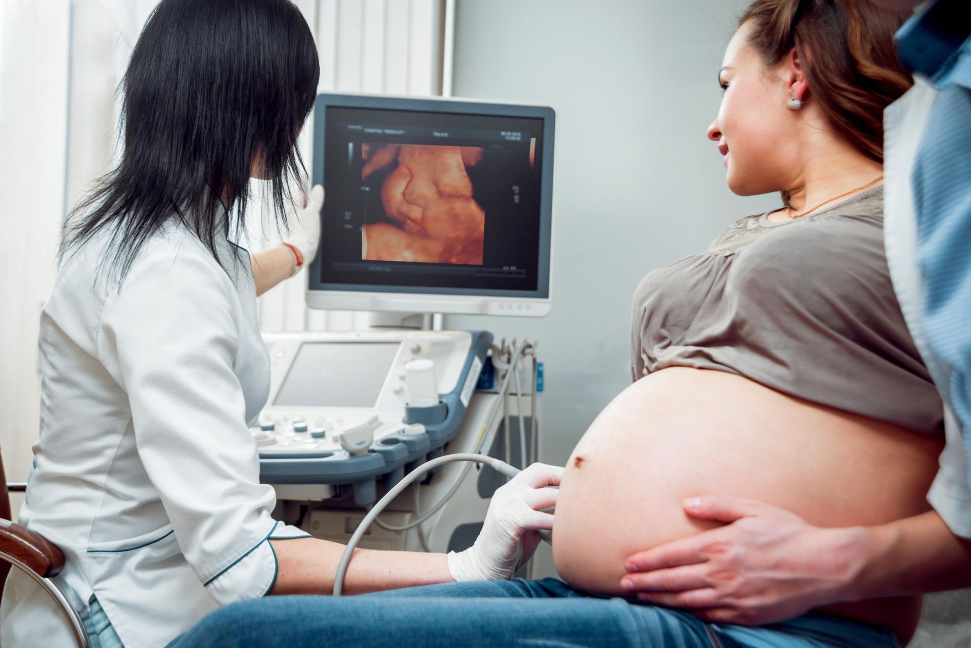 Pregnant woman undergoing ultrasound scan, doctor pointing at screen, partner's arm on belly, medical setting.
