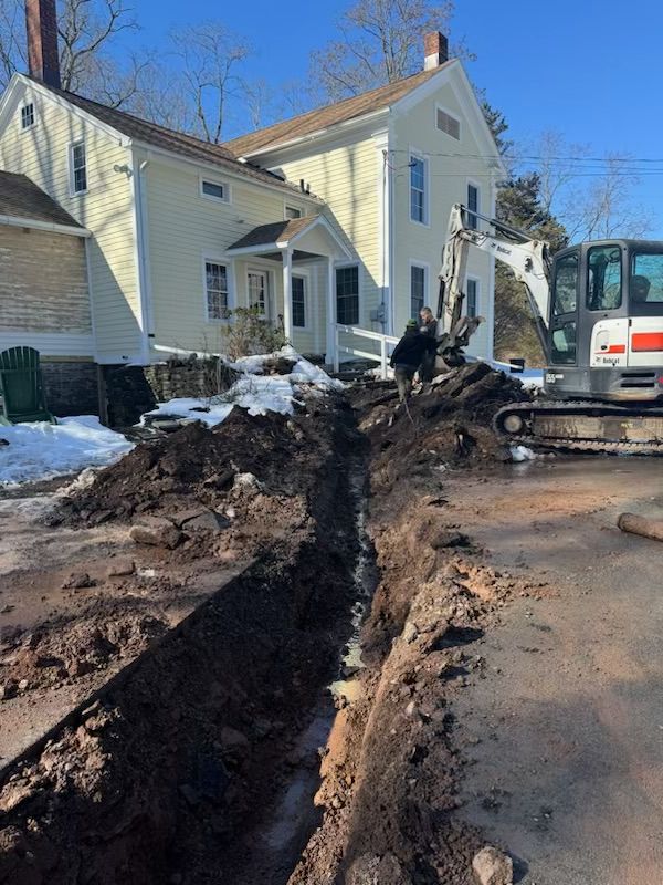 A worker in gloves and boots inserts a large suction hose into a rectangular septic tank opening in a grassy yard.