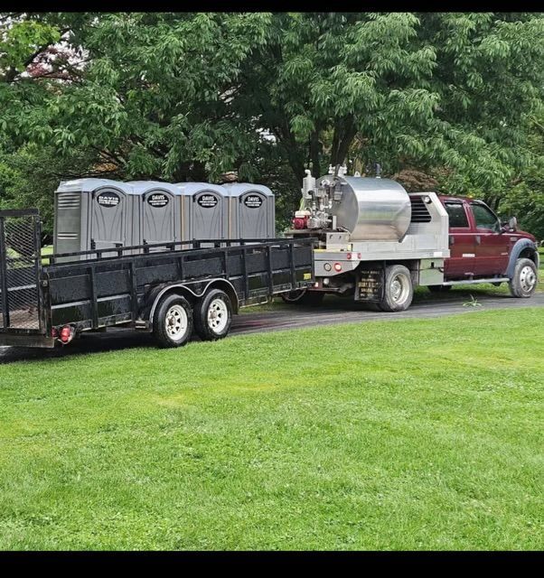 A dark red truck with a service tank hauling a trailer carrying four gray portable toilets on a grassy lawn.