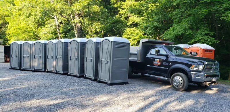 A black truck parked on a gravel lot next to a line of grey portable toilets in a wooded area.