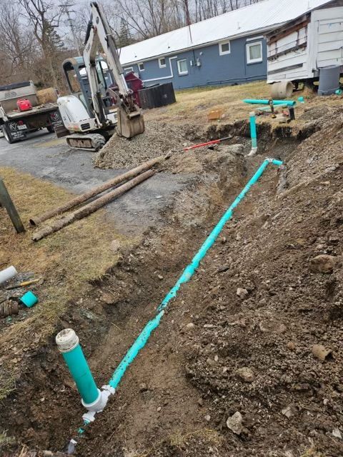 Excavated trench with green PVC piping and an upright pipe section near a small excavator and a blue house.