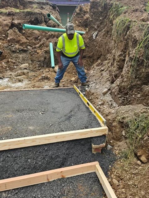 A worker in a high-visibility yellow vest uses a yellow level on wooden formwork in a gravel-filled construction trench.