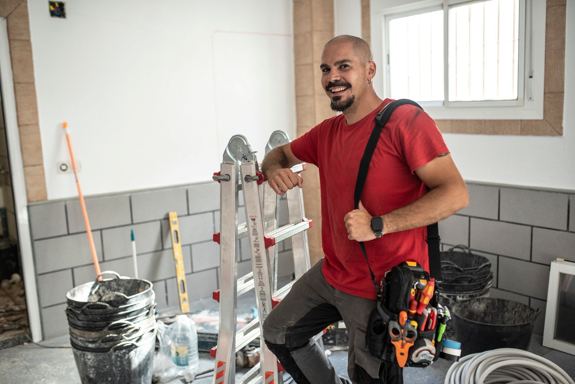 A man in a red shirt smiling with a tool belt by a ladder in a bathroom under renovation