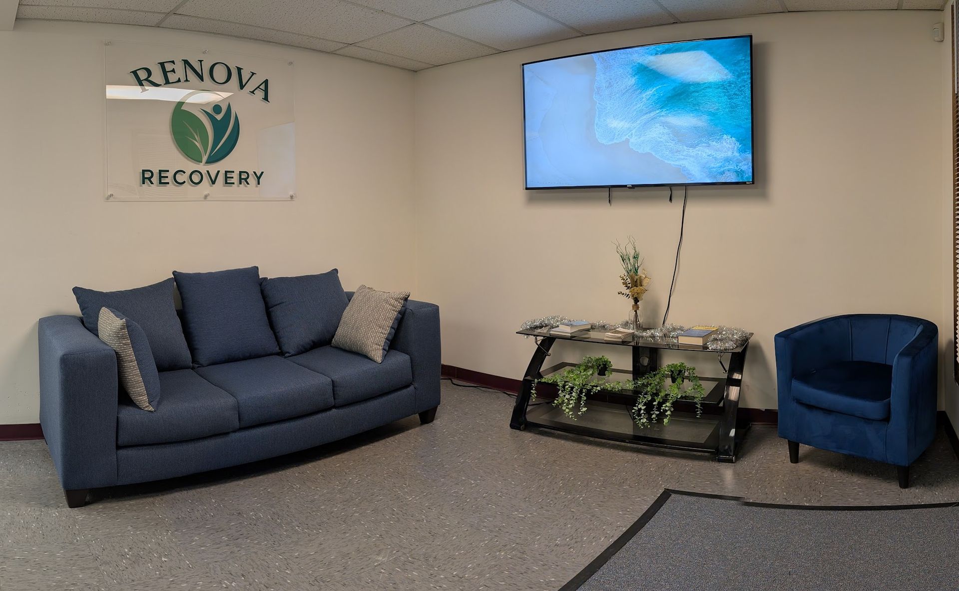 A waiting room with a blue sofa, a separate blue chair, and a wall-mounted TV showing a nature scene at Renova Recovery.