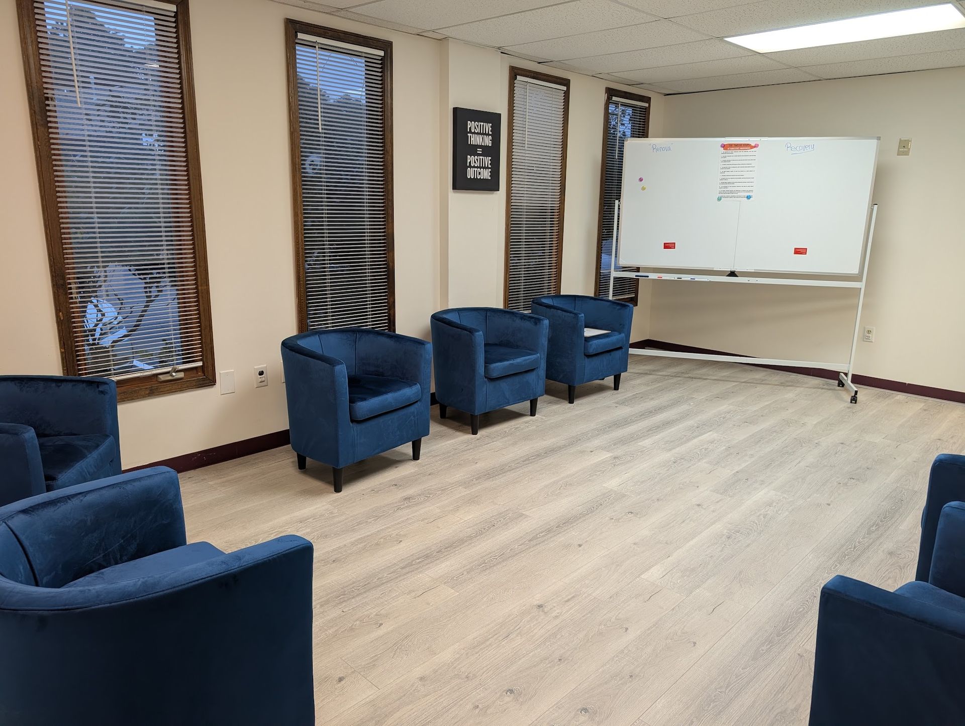 A meeting room with wood-look flooring, several blue armchairs arranged in a circle, and a rolling white board.