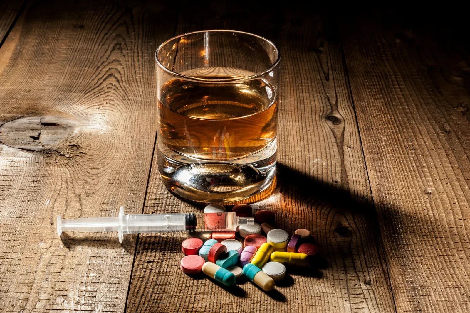 Whiskey glass beside syringe and scattered pills on a wooden table, suggesting substance abuse