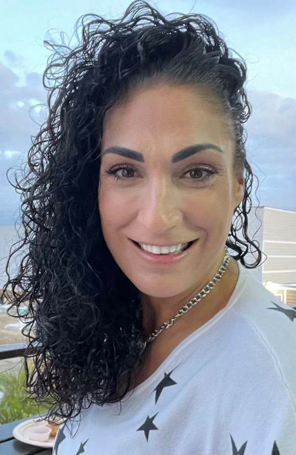 A smiling person with long hair wearing a black button-up shirt, posing with a hand near their neck by a window.