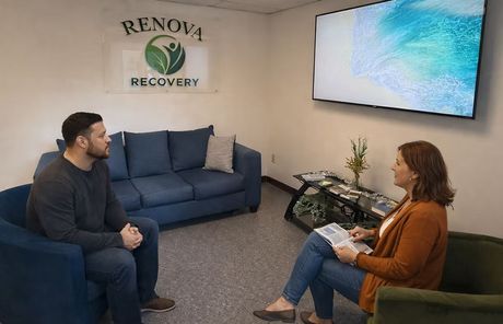 A counselor and client sit in chairs facing each other in an office with a blue couch and a Renova Recovery logo.