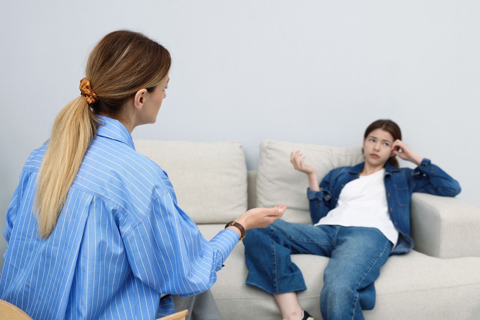 A person in a blue shirt speaks to a person sitting on a sofa, who appears disengaged or thoughtful.