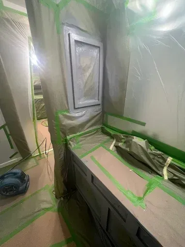 Room being prepped for painting: walls and furniture covered in plastic and green tape.