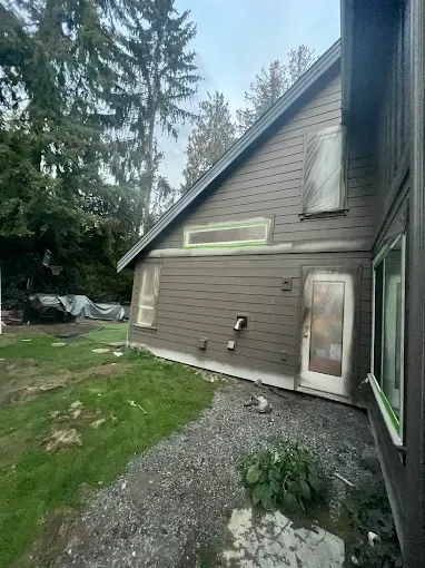 A brown house under renovation with green tape around windows and door, in a yard with gravel and grass.