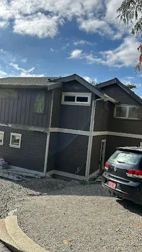 Dark brown house with white trim, parked car, gravel driveway, blue sky with clouds.