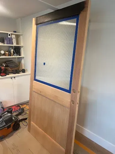 Wooden door being prepped for painting with blue tape around glass, tools on floor.