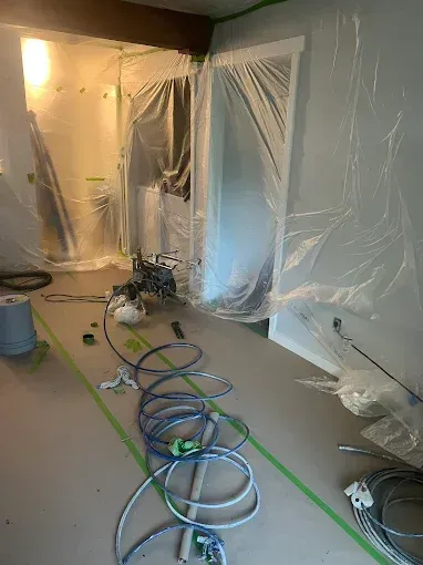 Room undergoing renovation, plastic sheeting covering doorways and surfaces, exposed tools and hoses on the floor.