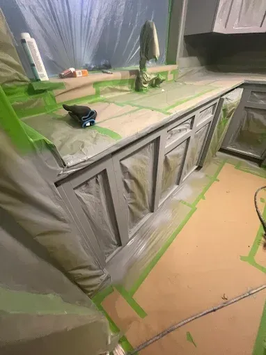 Kitchen cabinets being painted gray, with countertops and surrounding areas masked off with tape and plastic.