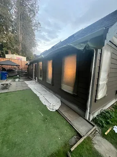 Brown house exterior with covered windows, and green grass yard under cloudy sky.