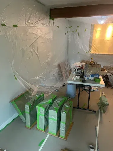 Room covered in plastic sheeting for painting. Green-taped boxes on the floor, a table with supplies.