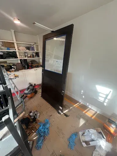 A dark stained door leaning in a room with messy flooring and white walls; work in progress.