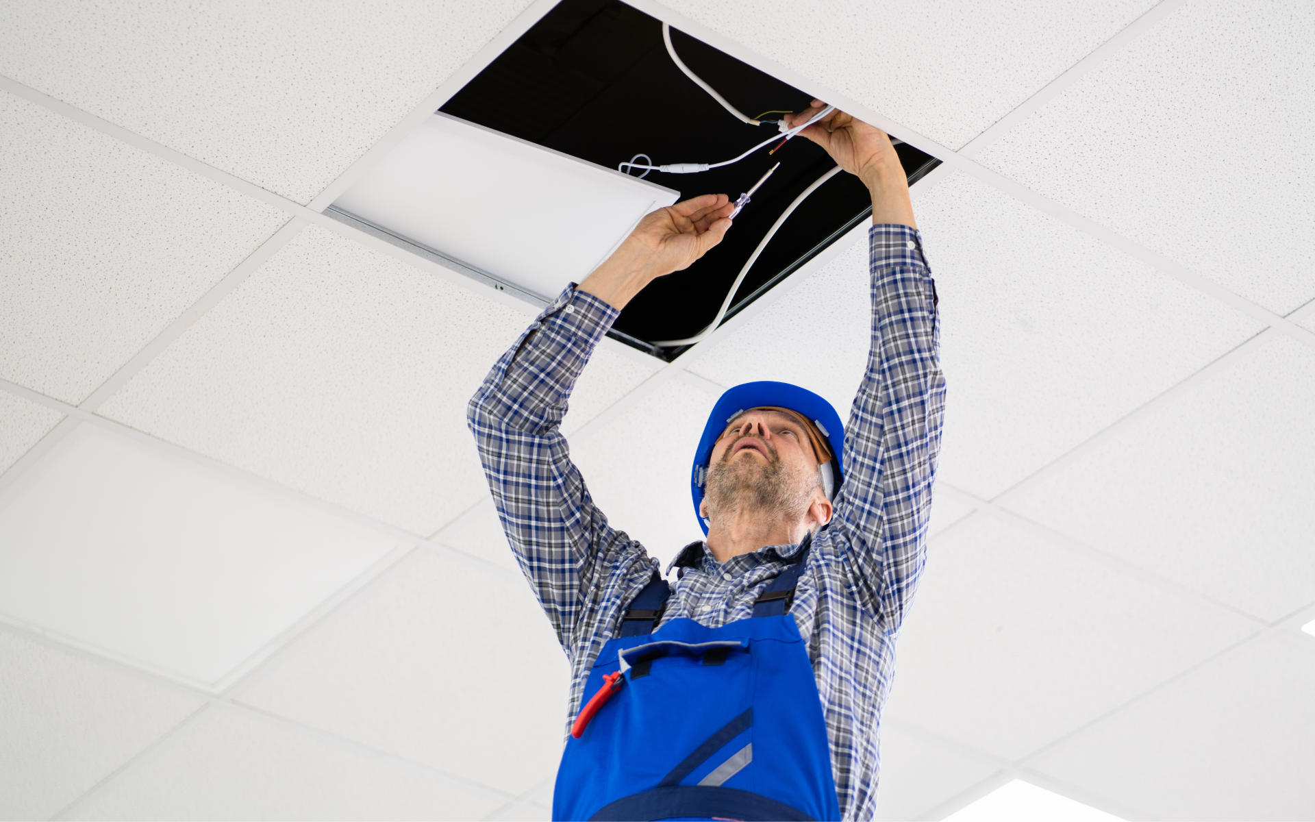 Electrician in blue coveralls and hard hat working on ceiling wiring.