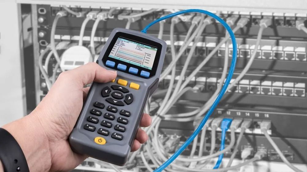 Hand holding a network cable tester in front of a network server with blue cable connected.