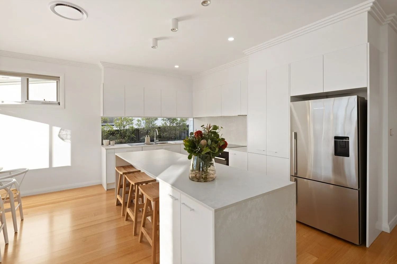Bright white kitchen with island, stainless steel fridge, wooden floors, and stools.