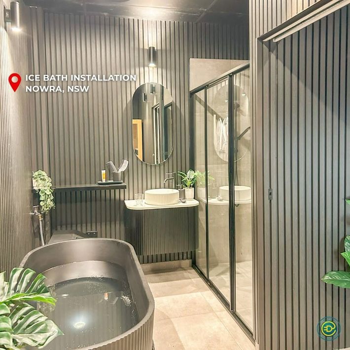 Dark modern bathroom with ice bath installation. Vertical gray paneling, black framed shower, oval mirror, and plants.