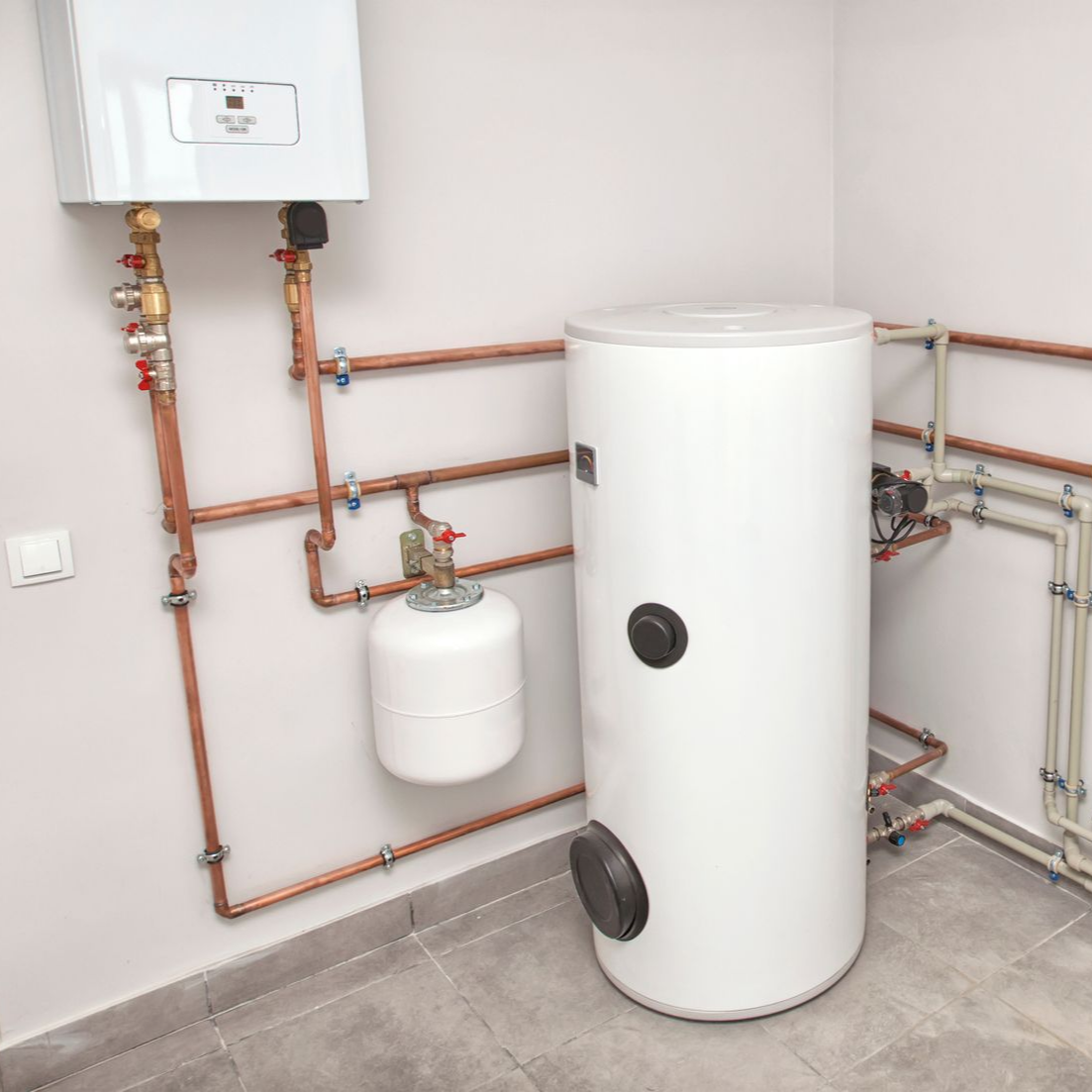 A white wall-mounted gas boiler connected to a large white cylindrical water heater with copper pipes in a tiled room.