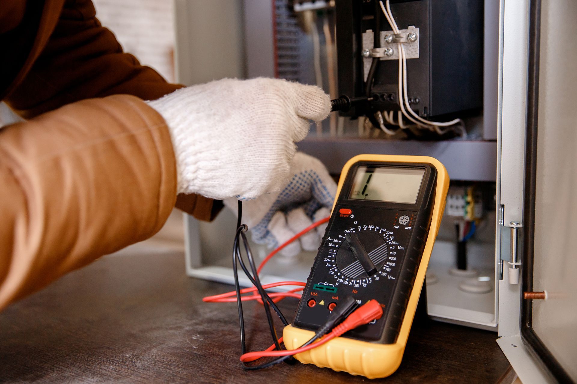 A person in white gloves uses a yellow digital multimeter to test electrical wires inside an open control panel.