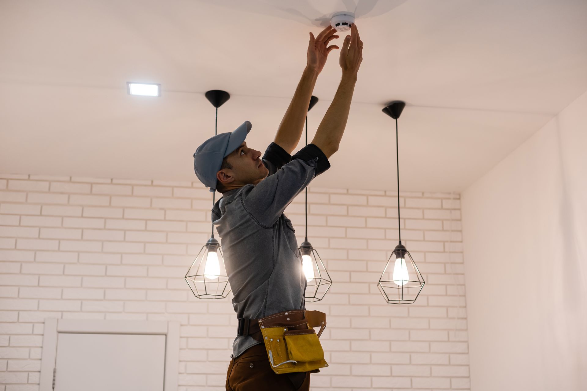 A person in a tool belt and cap stands on a ladder, reaching up to install a smoke detector on a white ceiling.