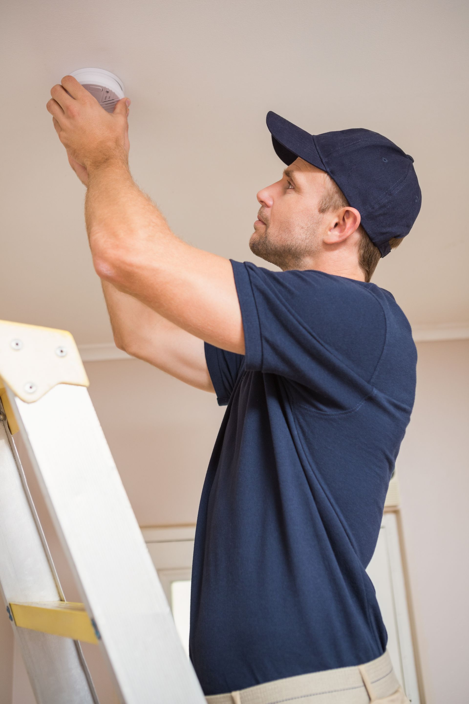 A person in a blue shirt and cap stands on a ladder, installing a white smoke detector onto a ceiling.