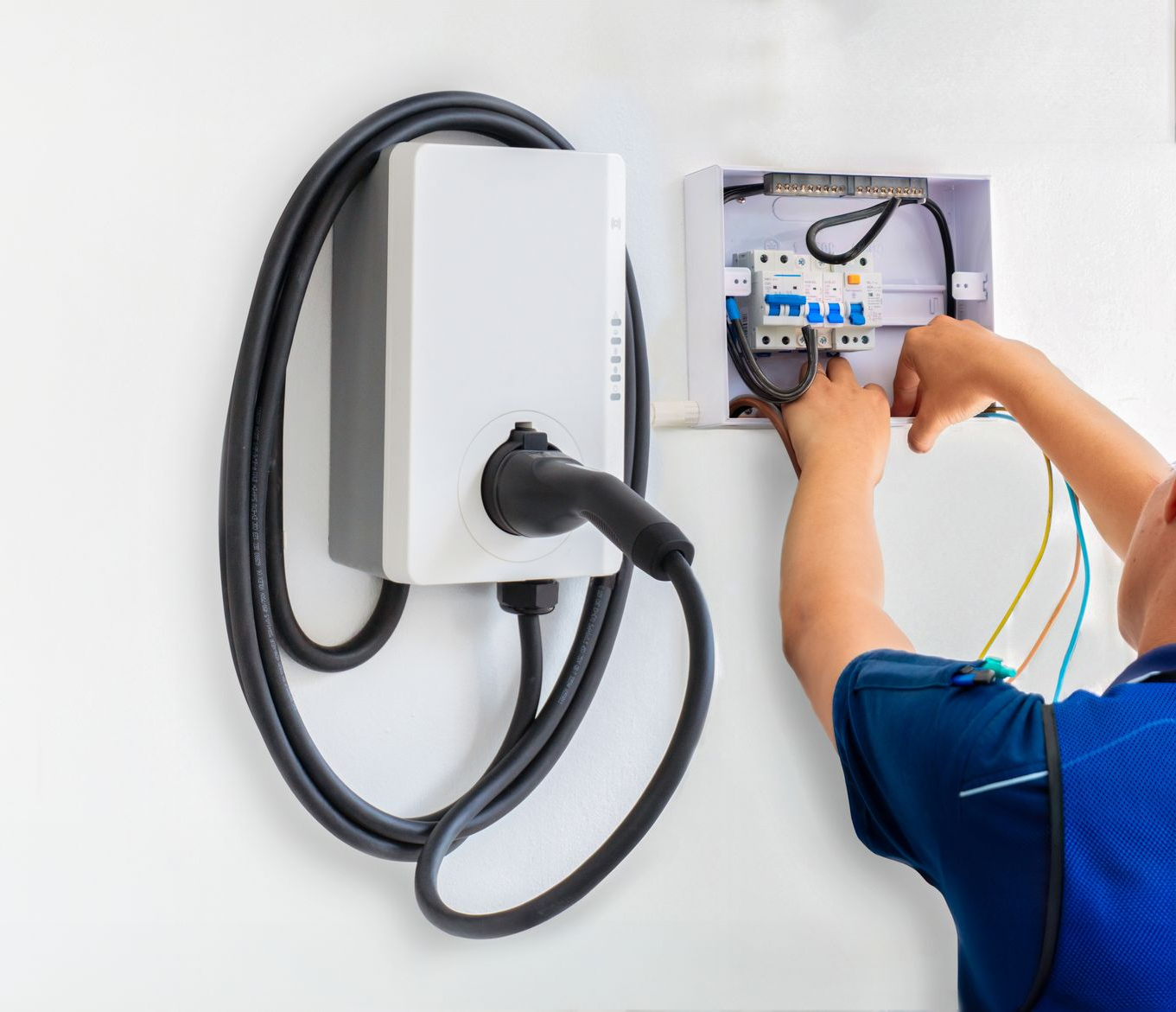 Electrician in a blue vest installing a wall-mounted electric vehicle charger and its circuit breaker box.