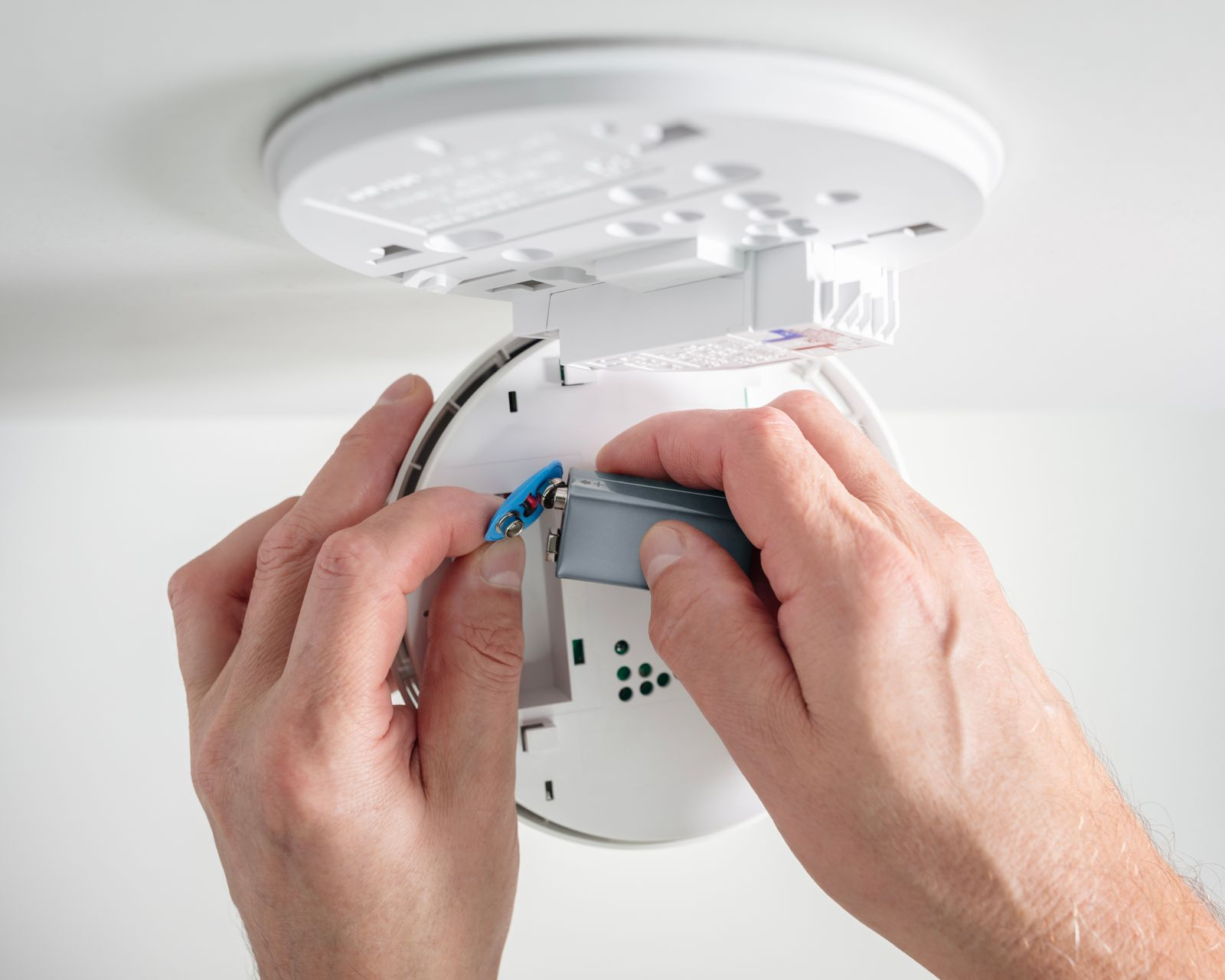 Hands removing or replacing a 9V battery in a white smoke detector mounted on a white ceiling.