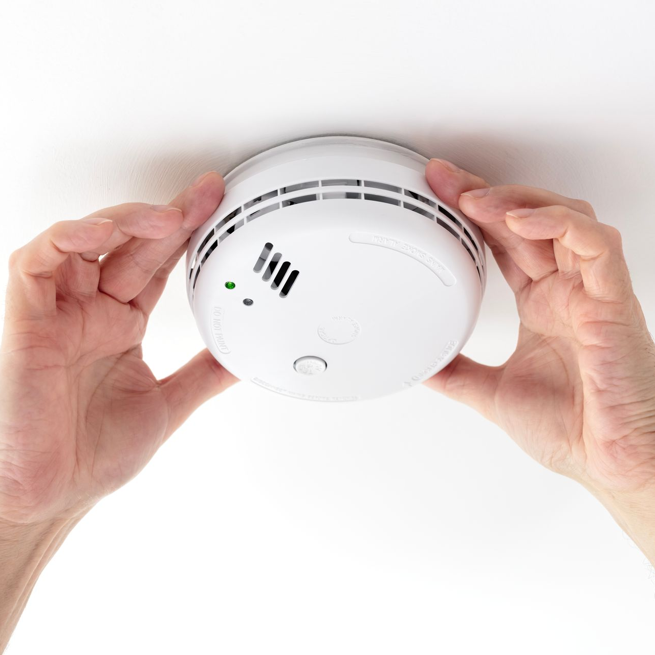 Hands installing or removing a white smoke detector from a white ceiling.