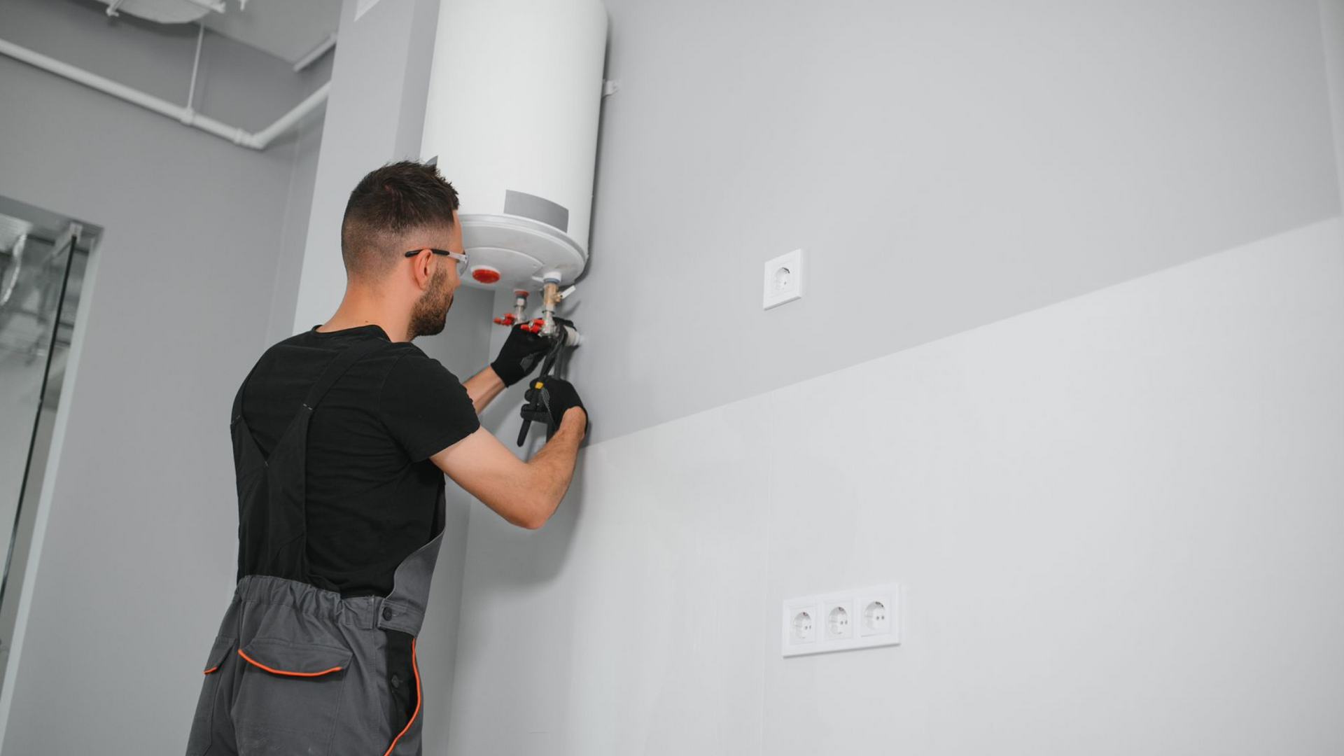 A person in work coveralls and gloves repairing pipes under a wall-mounted electric water heater in a room.