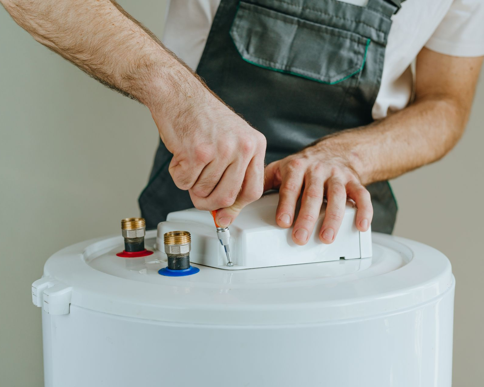 A person in work coveralls uses a screwdriver to attach a white cover to the top of a cylindrical water heater.
