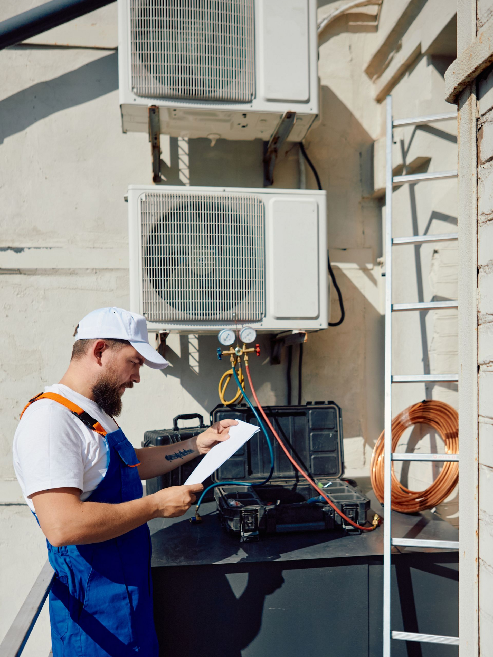 A technician in blue overalls and a white cap examines documents while working on outdoor air conditioning units.